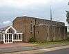 A bulky, plain brown brick building. A flat-roofed projection on the near side has four evenly-spaced ground-to-roof windows with concrete details; the main part of the building behind it has six evenly-spaced windows in concrete surrounds, the leftmost of which is much taller than the others. A tall, very thin green metal spire sits on the right side of the roof. A modern porch with white cladding is attached to the left side of the building.