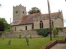 A stone church with red tiled roofs seen from the south. Extending from the chancel on the right is a battlemented chapel and, to the left, the tower is also battlemented