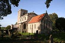 A stone church seeen from the southeast. The chancel has a red tiled roof, the larger nave with clerestory has a battlemented parapet, and the tower has a plain parapet