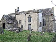 A small stone church seen from the southeast with a porch to the left, a protruding rendered south aisle in the middle, and a short chancel to the right. On the west gable is a bellcote, and in front of the church is a churchyard cross