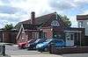 A long, low building of brick with a tiled roof, with extensions on the near side (next to a car park) and at the front. Behind the flat-roofed entrance porch is a semicircular window and a white crucifix. The nearer of the extensions on the left has a small chimney-stack.