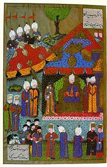 A woman holding a baby in her arms stands before a man who wears a turban and sits on a throne in a tent which is surrounded by dozens of people