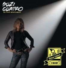 The front cover of Suzi Quatro's box set In the Spotlight Deluxe Edition. Quatro is standing up with her hands on her hips. A white spotlight shines on her from the top right, partly illuminating a black background. She is wearing a black leather jacket (over a black top) and light blue jeans with horizontal holes ripped in them. She has two black leather belts - a plain one, plus one decorated with yellow studs and yellow chains. In the bottom right there is a yellow rectanglular logo with the words "Deluxe Edition" and "2 CD Set".