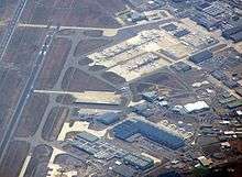 View from the air. Runway to the left and bottom. To the right long buildings and lots of aircraft.
