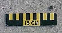 A ball of tar about 1 cm (0 in) wide on sand above a 15 cm (6 in) scale