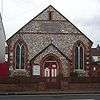 Front view of a tiny, plain chapel of irregular flintwork and red brick dressings. A dark stone plaque below a slit window near the roofline reads "WESLEYAN CHAPEL". An entrance porch is flanked by two wide, low-set windows with pointed arches.