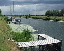 A broad, flat artificial cut with dark water and bright green raised banks. A patch of green algae stains the water. Several modern yachts are moored on wooden jetties spaced along the bank