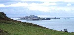 Brown-coloured islands of varying elevations lie in a calm sea beneath cloudy skies beyond a grassy slope.