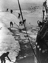 Men with digging tools removing ice surrounding the ship's hull, creating an icy pool of water