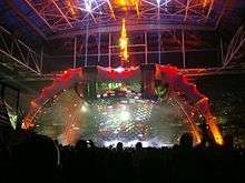 A tour stage; four large legs curve up above the stage and hold a video screen which is extended down to the band. The legs are lit up in red at the top and orange at the bottom. The video screen has multi-coloured lights flashing on it. The audience surrounds the stage on all sides.