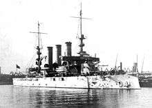 A white battleship with three smokestacks and two tall masts sitting in port.