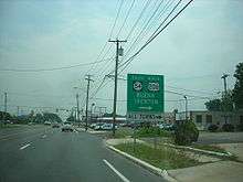 A four lane undivided road in a business area approaching a traffic light. A sign on the right side of the road reads south Route 54 north U.S. Route 206 Buena Trenton right all turns right