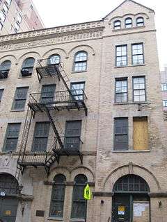 The right half of an old, sturdy-looking 4-story, 5-bay building, faced with partly soiled beige brick and architectural decorations. The right bay has a gabled fifth floor with 3 small windows. A fire escape runs down the front. At street level 1½ large doorways are visible. One of the 20 or so windows is boarded, another is painted black.