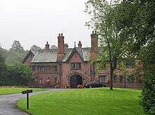 A two-storey house including an archway under a gable and another gable with half-timbering. In front of it is an area of grass and trees.