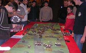 Several men surround a table, on which lies a small scale model of an undulating ground. Many small figures of monsters and medieval warriors and siege weapons are placed on that terrain. Dice are scattered among the figures.