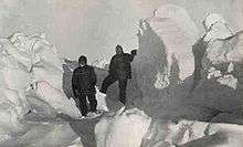 Two men in heavy clothing stand surrounded by mounds of ice which extend well above the height of their heads