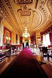 A photograph of a large room with a long red carpet stretching through the middle of it and windows on the right hand side. Furniture fills both sides of the room. The ceiling contains ornate plasterwork and a chandelier hangs down from the middle of the picture.